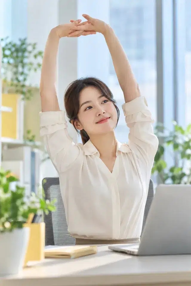 사무실 책상 앞에서 두 팔을 머리 위로 뻗어 스트레칭하는 직장인