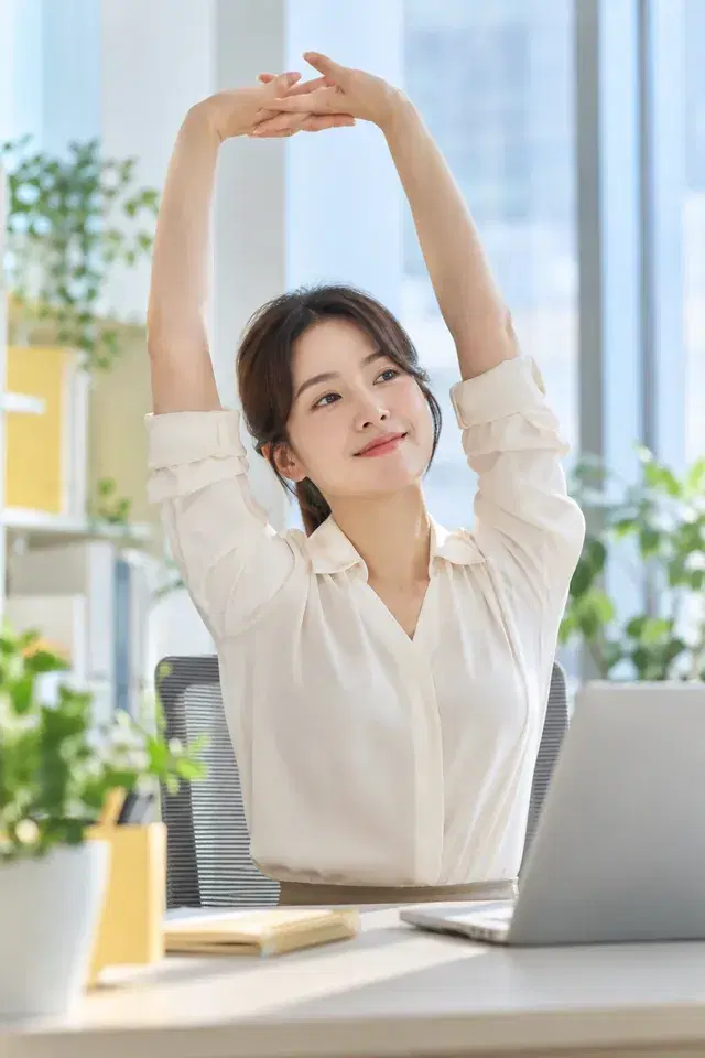 사무실 책상 앞에서 두 팔을 머리 위로 뻗어 스트레칭하는 직장인
