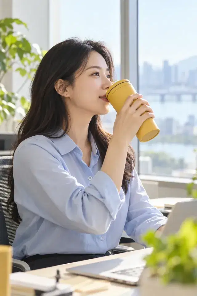 사무실 창가 책상에서 개인 텀블러로 음료를 마시는 직장인